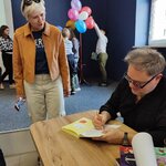 Osoba siedząca przy stole podpisuje książkę. Obok stoi dorosła osoba. W tle balony na sce.jpg