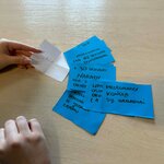 Zdjęcie przedstawia dłonie osoby trzymającej i układającej małe, kolorowe kartki z odręcznymi notatkami na jasnym blacie stołu. Na kartkach widoczne są krótkie zapiski w języku polskim.