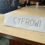 Na stole stoi kartka z napisem „CYFROWI”, a w tle widać osoby przy wspólnym stole w sali szkolnej lub bibliotece.