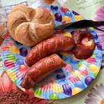 Na zdjęciu widać kolorowy talerzyk, a na nim 2 pieczone kiełbaski, nadgryzioną bułkę, kleks ketchupu oraz nóż.
