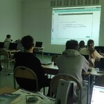 Uczniowie siedzą w pracowni komputerowej i patrzą na ekran z wyświetloną prezentacją. Na ławkach stoją laptopy, a obraz jest rzutowany na ścianę.
