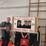 Czworo uczniów i opiekun stoją przed dekoracją świetlną w sali gimnastycznej. Trzymają ramę informacyjną dotyczącą zawodów oraz torbę pierwszej pomocy.