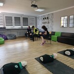 Sala szkolna z manekinami do resuscytacji ułożonymi na matach. Kilku uczniów siedzi na krzesłach i ćwiczy pierwszą pomoc przy użyciu sprzętu treningowego.
