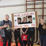 Grupa uczniów i opiekun stoją w sali gimnastycznej, trzymając dużą ramę z napisem „Zawody w udzielaniu pierwszej pomocy VIII LO”. Jedna osoba trzyma czerwoną torbę medyczną.