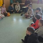 zdjęcie przedstawia część sali lekcyjnej z usadzonymi na podłodze uczniami, wśród nich na krzesełku siedzi nauczycielka z książką na kolanach. Na wprost duże okno zasłonięte jasnozieloną roletą. Po lewej stronie duża szafa i kilka mniejszych żółtych i zielonych drzwiczek od szafek.
