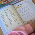 Zbliżenie na szkolną ławkę: otwarty zeszyt ćwiczeń z listą emocji i uzupełnionymi zdaniami, obok piórnik i podręczniki. Dłoń dziecka opiera się na szkolnym blacie.