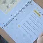 Widok z góry na szkolną ławkę: otwarty zeszyt ćwiczeń z listą emocji oraz zadaniem do uzupełnienia zdań, zapisany odręcznie. Po obu stronach zeszytu widoczne dłonie dziecka, a w tle drewniany blat ławki i fragment zielonego krzesła.