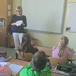 zajęcia w klasie szkolnej: osoba prowadząca stoi przy tablicy interaktywnej i trzyma karty z rysunkami, a uczniowie siedzą w ławkach i słuchają. Na ścianie widoczny godło państwowe, w sali znajdują się szafki, przybory szkolne i zeszyty na ławkach.