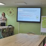 widok części sali lekcyjnej ze stolikami i postaciami uczestników od tyłu. W centralnej części ekran multimedialny z prezentacją, po lewej stronie kobieta, mediator sądowa, zwracająca się do uczestników, po prawej kobieta prowadząca spotkanie. Przy ekranie multimedialnym 2 flipcharty jeden za drugim, na pierwszym flipcharcie żółte karteczki samoprzylepne.