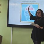 w centralnej części ekran multimedialny z prezentacją, po lewej stronie kobieta, mediator sądowa patrząca na ekran, centralnie, na tle ekranu, kobieta z uniesioną ręką wskazującą treści prezentacji. Przy prawej krawędzi flipchart z żółtymi karteczkami samoprzylepnymi.