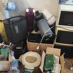 Na zdjęciu widać stertę różnych urządzeń elektrycznych i elektronicznych ustawionych pod ścianą.  Są tam m.in. mikrofalówki,  wentylatory, przewody, elementy elektroniczne, pojemniki i sprzęt AGD. 