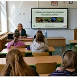 Na zdjęciu widać panią  prowadzącą zajęcia edukacyjne w klasie szkolnej. Uczniowie siedzą w ławkach i słuchają prezentacji wyświetlanej na tablicy multimedialnej.  