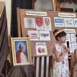 Dwie dziewczynki stoją na scenie z mikrofonami obok portret Jana Pawła II_ tablica „Patron.jpg