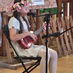 Dziecko siedzi na krześle i gra na ukulele przy mikrofonie za nim tablica „Patronat medial.jpg