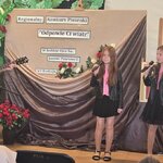 Na scenie dorosła osoba gra na gitarze_ obok dwoje dzieci śpiewa do mikrofonów w tle dekor.jpg