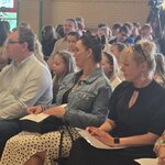 Sala z ustawionymi rzędami krzeseł liczna publiczność siedzi i słucha_ w tle okna_ rośliny.jpg