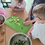 Troje dzieci przy stoliku kroi warzywa nożem na zielonej desce widoczne miski_ obierki cuk (1).jpg