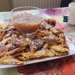 Na talerzu są kawałki smażonego placka, posypane cukrem pudrem oraz pucharek z konfiturą. Obok stoi kubek z herbatą.