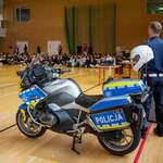 W sali gimnastycznej stoi policyjny motocykl z widocznym napisem „Policja”, obok znajduje się stanowisko informacyjne oraz funkcjonariusz trzymający kask. W tle siedzi duża grupa uczniów, którzy obserwują prezentację, a na ścianach widoczne są drabinki i sprzęt sportowy.