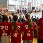 W sali gimnastycznej duża grupa uczniów siedzi na podłodze i ogląda prezentację; na pierwszym planie stoi pięć osób w czerwonych koszulkach z napisem „SP 12 to MY!”. W tle widoczne są stoiska informacyjne, ekran projekcyjny, policyjny motocykl oraz materiały promujące bezpieczeństwo.