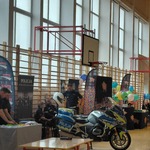 Na sali gimnastycznej ustawione jest stoisko Policji z materiałami informacyjnymi oraz policyjny motocykl. Obok stoją plansze, flagi promocyjne i kolorowe balony, a kilka osób obsługuje stanowisko. W tle widoczne drabinki gimnastyczne i kosze do koszykówki.