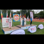 Trzy młode zawodniczki z dyplomami pozują na plenerowym podium podczas zawodów Szkolnego Związku Sportowego.png