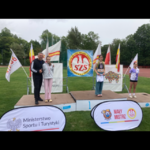 Trzy młode zawodniczki i mężczyzna stoją na podium podczas ceremonii dekoracji.png