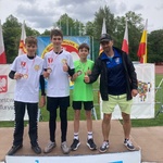 Trzej młodzi zawodnicy z medalami oraz dorosły mężczyzna stoją na plenerowym podium podczas dekoracji.jpeg