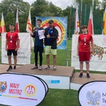 Trzej młodzi zawodnicy pokazujący medale oraz mężczyzna stoją wspólnie na najwyższym stopniu plenerowego podium.jpeg