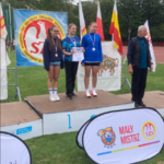 Trzy młode zawodniczki na plenerowym podium obok mężczyzny widoczne są flagi_ baner Szkolnego Związku Sportowego.png