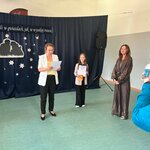 Podczas uroczystego podsumowania XXVIII Wojewódzkiego Konkursu Recytatorskiego „Świat malowany poezją Wiesława Kazaneckiego” uczennica klasy 5b Kornelia G. stoi z dyplomem i nagrodą. W tle widoczna jest dekoracja konkursowa z motywem gwiazd, a organizatorzy ogłaszają wyniki wydarzenia.