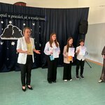 W sali szkolnej odbywa się uroczystość wręczenia nagród w konkursie recytatorskim. Na tle granatowej kurtyny ozdobionej papierowymi gwiazdami i napisem „Będziemy szeptali w gwiazdach, jak w wysokiej trawie” jedna osoba przemawia do zgromadzonych. Obok stoją uczniowie trzymający dyplomy i upominki. Wśród nagrodzonych znajduje się uczennica klasy 5b, Kornelia G., która zdobyła I miejsce w XXVIII Wojewódzkim Konkursie Recytatorskim „Świat malowany poezją Wiesława Kazaneckiego”, organizowanym przez Szkołę Podstawową nr 38 w Białymstoku. W tle widoczny jest głośnik oraz dekoracja z sylwetkami postaci na tle gwiazd.