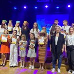 Grupa uczniów stoi na scenie po zakończeniu konkursu wokalnego, trzymając dyplomy i statuetki. Uczestnicy ustawieni są w kilku rzędach, a obok nich stoją osoby dorosłe. Scena oświetlona jest niebieskim światłem, a w tle widoczny jest ekran z napisem dotyczącym konkursu piosenki.