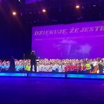 Duży chór dzieci stoi na scenie podczas koncertu w sali widowiskowej, a przed nim osoba dyrygująca. Scena oświetlona jest fioletowym światłem, a w tle widoczny jest napis „Dziękuję, że jesteś! Koncert o rodzinie, przyjaźni i marzeniach”. W chórze uczestnicy ubrani są w różne kolorowe stroje, a po prawej stronie sceny widoczni są muzycy z instrumentami. 