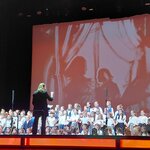 Chór dzieci stoi na scenie podczas koncertu, a przed nim osoba dyryguje zespołem. Na dużym ekranie w tle wyświetlana jest projekcja w ciepłych, czerwonych barwach przedstawiająca sylwetki postaci.  