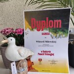 Dyplom nagrody w konkursie piosenki wakacyjnej dla Wiktorii Milewskiej, ustawiony obok dekoracyjnej figurki ptaka i różowych kwiatów