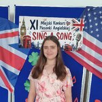 Uczennica stoi na tle dekoracji z flagami Wielkiej Brytanii i USA oraz napisem „XI Miejski Konkurs Piosenki Angielskiej Sing a Song”