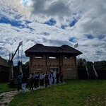 Grupa uczniów stoi przed drewnianą rekonstrukcją średniowiecznego grodu. Nad budowlą rozciąga się zachmurzone niebo z prześwitującym słońcem.
