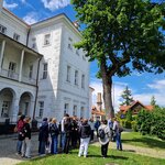 Uczniowie uczestniczą w zwiedzaniu przy białym zabytkowym budynku otoczonym zielenią. W tle widoczne jest duże drzewo i słoneczna pogoda.