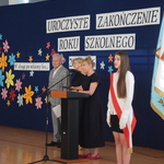 Uroczyste zakończenie roku szkolnego – przemówienie przy mównicy, obok uczennica z biało‑czerwoną szarfą.