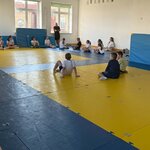 Grupa dzieci siedzi na matach w sali gimnastycznej podczas zajęć sportowych, a prowadzący stoi przy oknie i udziela instrukcji.