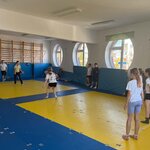 Dzieci uczestniczą w zajęciach ruchowych w jasnej sali gimnastycznej z niebiesko-żółtymi materacami i dużymi okrągłymi oknami.