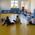 Dzieci siedzą w kręgu na matach w sali gimnastycznej, słuchając wskazówek prowadzącego stojącego przy oknie.