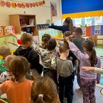 Grupa dzieci stoi w sali lekcyjnej i wykonuje wspólne ćwiczenie ruchowe, wyciągając ręce w jednym kierunku. W tle widoczne są szafki, dekoracje z jesiennych liści oraz plansze edukacyjne z nazwami polskich miast.