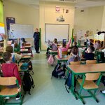 Dzieci siedzą w ławkach w sali lekcyjnej i uczestniczą w spotkaniu edukacyjnym prowadzonym przez funkcjonariuszkę policji. Kilkoro uczniów zgłasza się do odpowiedzi, unosząc ręce. W tle widoczne są tablica, pomoce dydaktyczne oraz wyposażenie klasy.