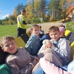 Na głównym planie zdjęcia widać troje uczniów, dwóch chłopców, którzy patrzą prosto przed siebie i jedną dziewczynkę, która opiera się rękoma. Dzieci siedzą w wiejskim wozie w kolorze żółto-czarnym. Na drugim planie widać mężczyznę w żółtej koszulce i czarnych spodniach, który idzie na tył wozu. W wozie jedzie także trójka dzieci, którzy są w dalszym planie. W tle widać budynek i zadrzewienie.