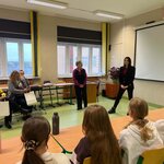 „Spotkanie w sali lekcyjnej: dwie kobiety stoją przed uczniami, a pozostałe uczestniczki wizyty siedzą przy szkolnych ławkach.”