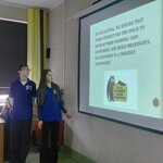 „Dwoje uczniów w koszulkach SP12 przedstawia po angielsku prezentację o szkole; na ekranie widnieje jej logo.”