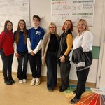 „Uczestniczki wizyty z nauczycielką i dwojgiem uczniów w koszulkach SP12 na szkolnym korytarzu, przy planszach matematycznych.”