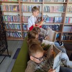 Uczniowie klasy 2c podczas wspólnego czytania książek w szkolnej bibliotece. Dzieci korzystają z kącika czytelniczego wyposażonego w fotele, kanapy i stolik.